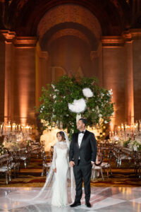 A bride and groom stand holding hands in an elegant, grand hall with high ceilings, ornate decor, and a large floral arrangement behind them. The bride wears a white lace gown and veil; the groom is in a black tuxedo.