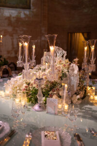 Elegant table setting with crystal candle holders, lit candles, white flowers, and a table number card. Soft, warm lighting creates a romantic and luxurious atmosphere.