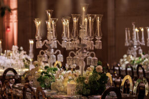 Elegant banquet table set with tall crystal candelabras, surrounded by gold chairs, white flowers, and lush greenery. Warm lighting creates a luxurious and festive atmosphere in a spacious hall.