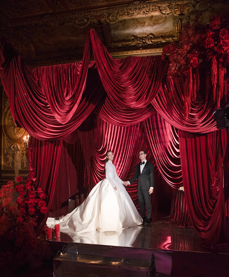 A bride in a white gown and a groom in a black tuxedo stand on a stage with dramatic red velvet drapes and red floral arrangements, under ornate gold decor. The scene is elegant and theatrical.